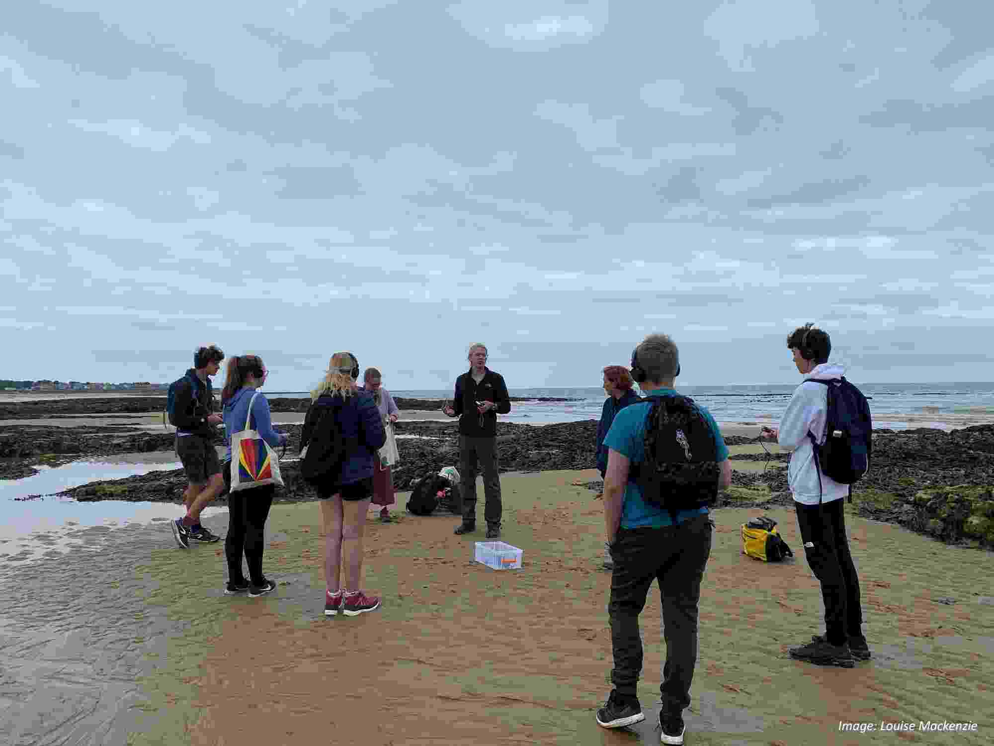 Eight people stand in a circle near to some rocks on the seashore